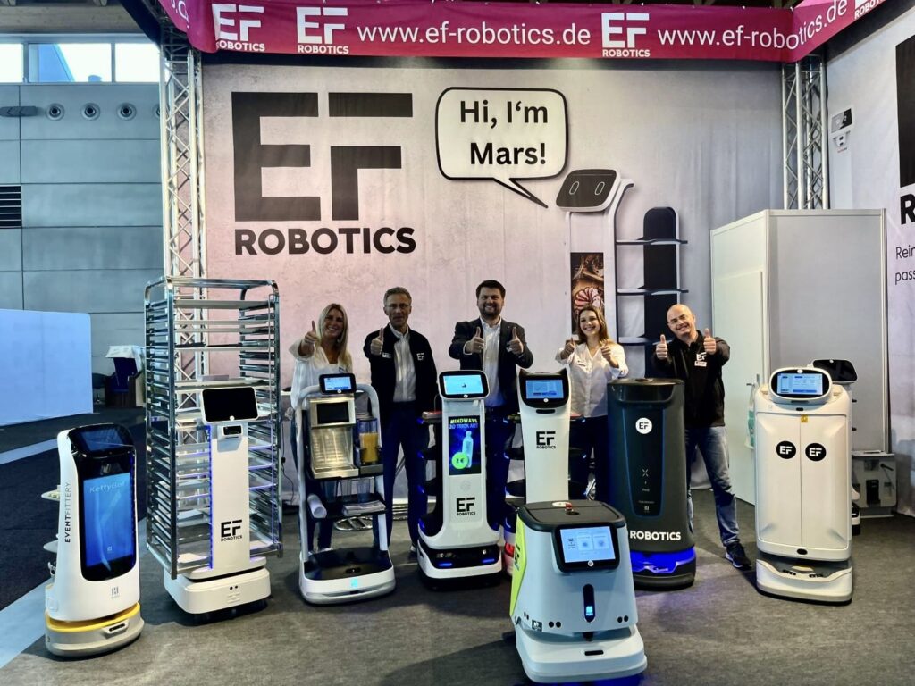 Das Team von EF Robotics grinst freundlich in die Kamera vor dem Stand von EF Robotics. So kann man neue Handelspartner empfangen. 